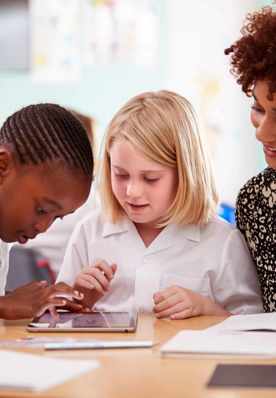 Female Teacher With Two Elementary School Pupils Wearing Uniform Using Digital Tablet At Desk